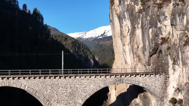 landwasser viaduct arches meet towering limestone cliff wall near filisur switzerland forested valley opens beyond snowcapped peak rises aerial drone footage clear blue sky mountain gorge scenery