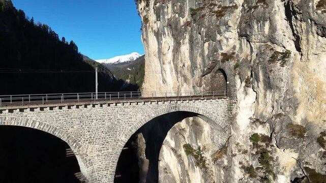railway line curves toward tunnel portal cut into limestone cliff on landwasser viaduct near filisur switzerland snowcapped peak rises beyond forested gorge aerial drone footage clear sky alpine