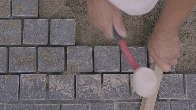 Top view of a builder leveling gray paving stones with a rubber mallet and wooden plank