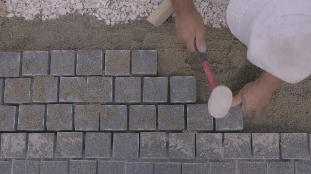 Top down view of a builder laying gray paving stones on sand with a rubber mallet