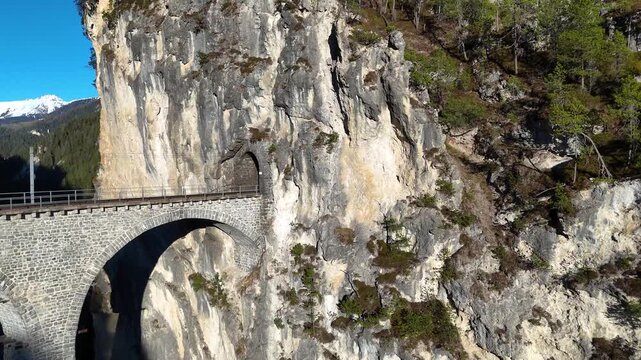 limestone cliff towers over landwasser viaduct curved arches near filisur switzerland tunnel portal cut rock face snowcapped peak forested slopes graubunden aerial drone footage clear sky scenery