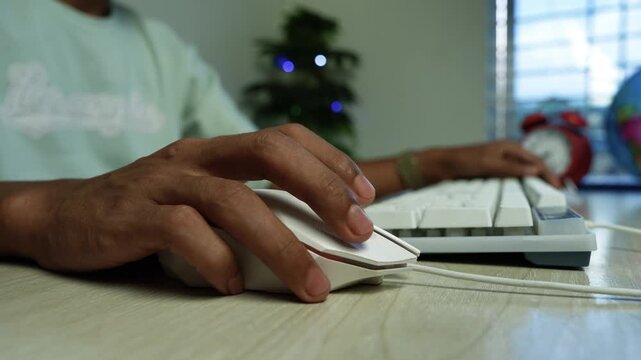 Person Using Mouse and Keyboard at Home Office, Close-up of a person using a computer mouse beside a keyboard at a desk, with a small decorated Christmas tree blurred in a home office background.