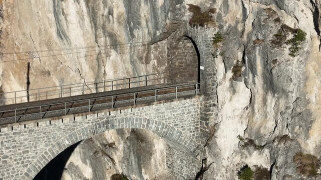 weathered rock face frames landwasser tunnel entrance stone archway curved viaduct track leads wires span gorge vegetation grips limestone ledges aerial drone footage filisur graubunden switzerland