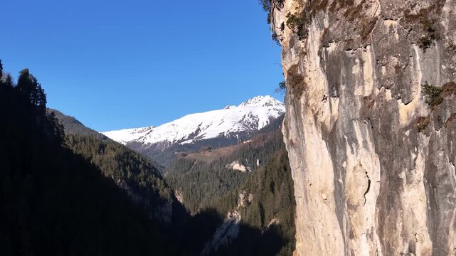 sheer limestone rock face towers over landwasser gorge near filisur switzerland snowcapped peak rises beyond pine forest mountainside aerial drone footage clear blue sky alpine scenery graubunden