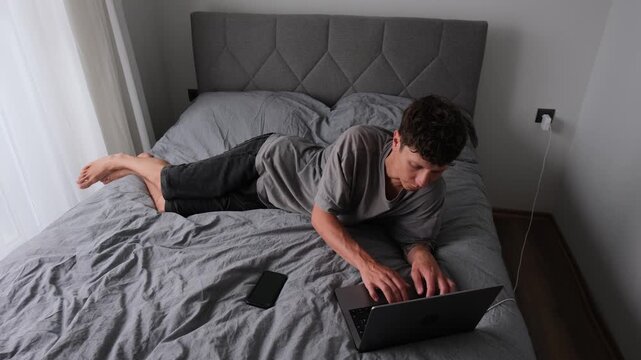 Man working on laptop in awkward position on bed at home, unhealthy posture causing back pain and physical discomfort, realistic student and remote work lifestyle in small apartment
