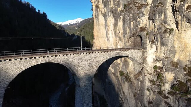 catenary wires trace landwasser viaduct arches toward tunnel opening limestone cliff near filisur switzerland snowcapped peak rises behind forested ridge aerial drone footage clear blue sky gorge