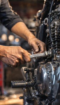 Mechanic hands tighten a bolt on a motorcycle engine using a wrench, showing worn hands and oily metal parts. The image focuses on manual repair work and mechanical detail.