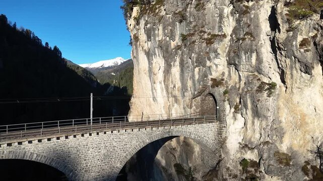 landwasser viaduct tunnel portal cuts limestone cliff filisur switzerland six arches curve forested gorge snowcapped peak catenary wires span track aerial drone footage clear sky alpine graubunden