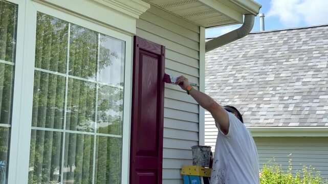 Handyman carefully painting the edge of a window shutter on a house
