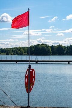 Red warning flag waving on a tall pole with an orange rescue buoy attached on a lake pier, representing water safety and no swimming alert under a blue cloudy sky.