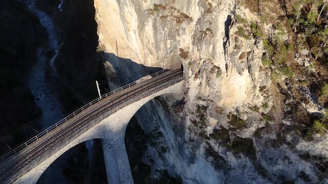 curved viaduct track descends tunnel portal etched limestone cliff filisur switzerland shadow crosses stone arches sparse pine clings ledges catenary wires steep angle aerial drone footage graubunden