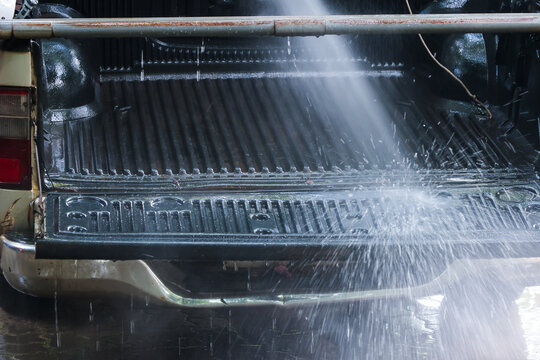 Pickup truck cargo bed receiving high pressure water spray during outdoor vehicle washing, creating bright droplets and clean reflective metal surface in sunlight