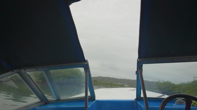 Blue boat cabin with open sunshades. Steady hand guides the steering wheel. Calm focus on the river journey. Lush green hills line the waterway. Overcast sky adds serene mood