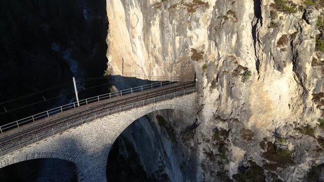 railway track angles past landwasser tunnel entrance limestone cliff filisur switzerland stone arches curve shadowed gorge sparse pine ledges catenary wires signal pole aerial drone footage graubunden