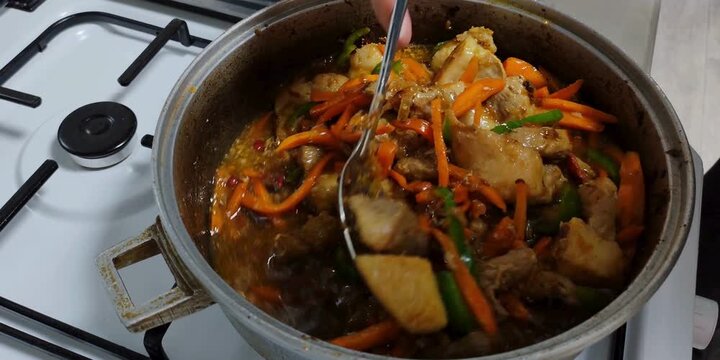 A chef prepares a hearty meal of cooked meat and sliced carrots stirred with a metal spoon inside a heavy metal pot on a kitchen stove to create a delicious homemade dinner for hungry families
