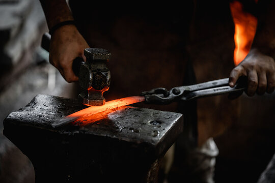 Blacksmith forging glowing hot iron on anvil with hammer and tongs in dimly lit workshop near blazing forge fire