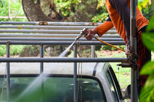 Worker pressure washing metal scaffold beside vehicle in leafy outdoor setting, wearing protective clothing while spraying water from elevated equipment