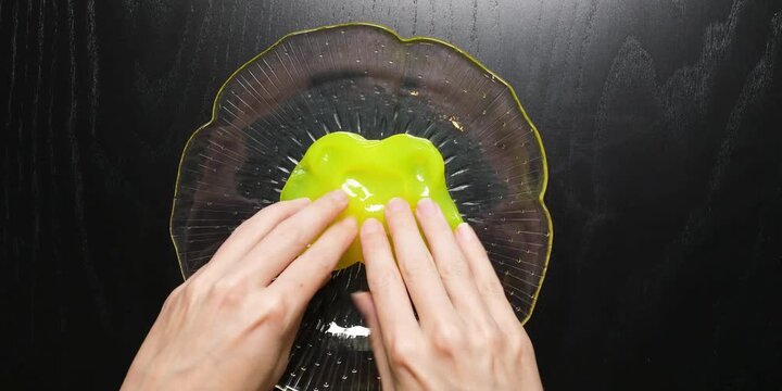 Hands pressing down on bright neon lime green slippery sensory putty placed atop a dark textured glass surface representing creative stress relief therapy play and fun tactile exploration for