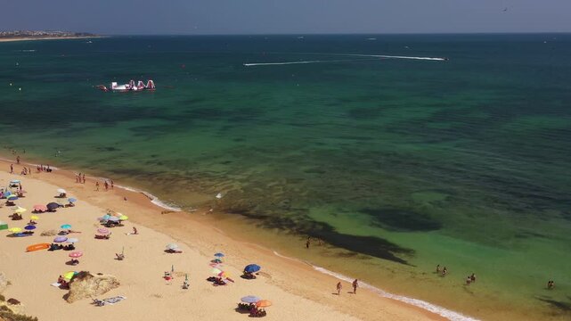 Drone flies above Armacao de Pera Beach in Algarve, revealing turquoise Atlantic waters, a sandy shoreline filled with beach umbrellas and swimmers enjoying a sunny summer day