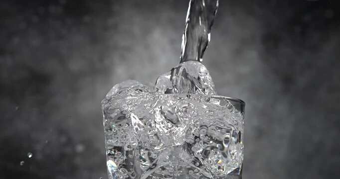 Water pouring into glass with ice cubes in slow motion on black background
