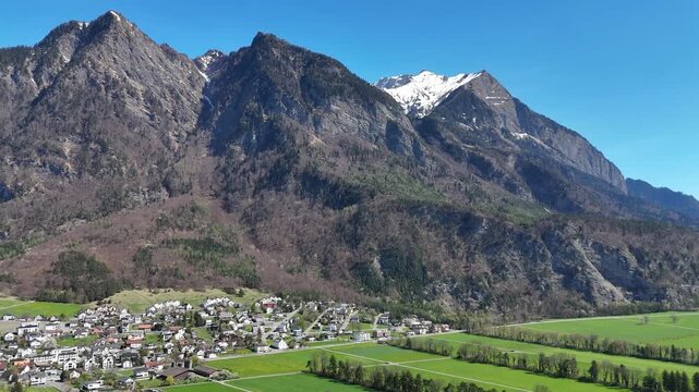 jagged alpine ridge rugged rock face towering small village houses green farmland fields forested slopes patchy snow liechtenstein rhine valley drone footage aerial view establishing shot clear sky