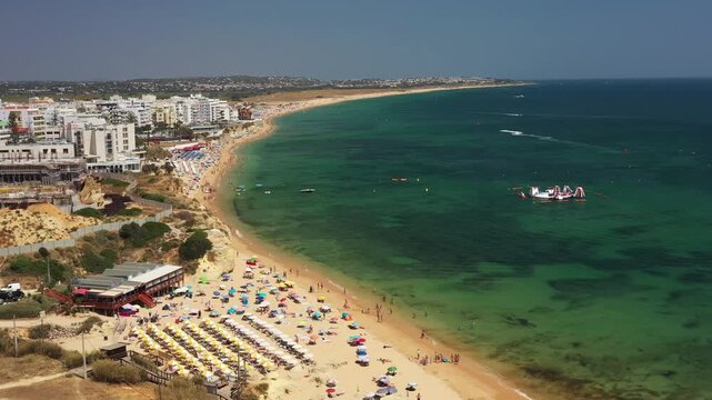 Drone follows the coastline of Armacao de Pera in Algarve, showcasing the resort town, golden sandy beach, turquoise Atlantic waters and vibrant summer atmosphere
