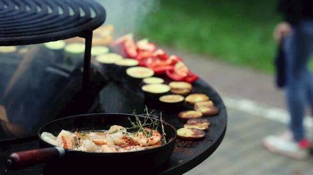 Shrimp in pan and vegetables grilling on outdoor fire pit grill. Fresh seafood with zucchini, pepper and tomato cooking on barbecue. People walking on park path in background.