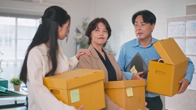 Group of fired Asian employees holding cardboard boxes with personal belongings, supporting and comforting each other after job dismissal in a modern office, representing workplace crisis.