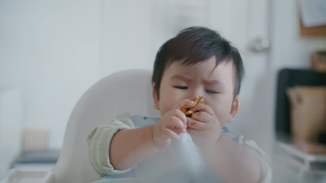 Asian baby boy eating boiled corn on high chair, BLW (baby-led weaning) concept, healthy solid food introducing to infant at home