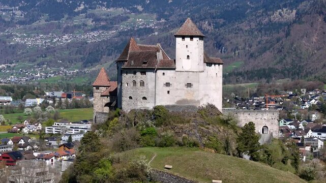 gatehouse stone walls gutenberg castle round tower forested mountain slope backdrop balzers town rooftops construction crane blossom trees aerial orbit drone footage liechtenstein heritage site