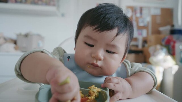 Asian baby boy eating boiled corn on high chair, BLW (baby-led weaning) concept, healthy solid food introducing to infant at home
