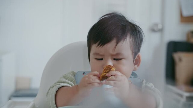 Asian baby boy eating boiled corn on high chair, BLW (baby-led weaning) concept, healthy solid food introducing to infant at home