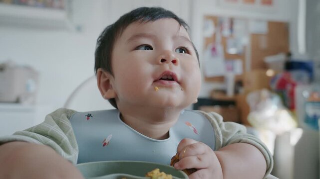Asian baby boy eating boiled corn on high chair, BLW (baby-led weaning) concept, healthy solid food introducing to infant at home
