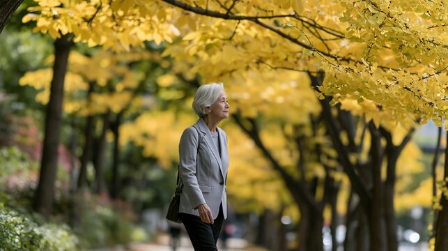 an elderly woman walking through a park. She wears a light gray blazer and dark pants, carrying a dark handbag. The womans short, white hair falls down her back, and she gazes upwards.