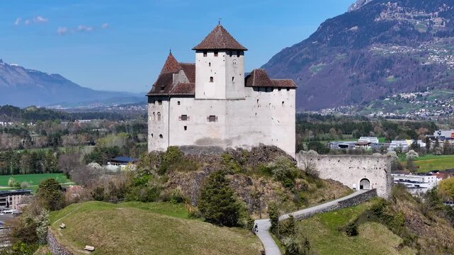 wide valley panorama gutenberg castle hilltop jagged snowcapped alpine ridge backdrop parish church nearby balzers rhine valley farmland town rooftops drone footage aerial orbit heritage site