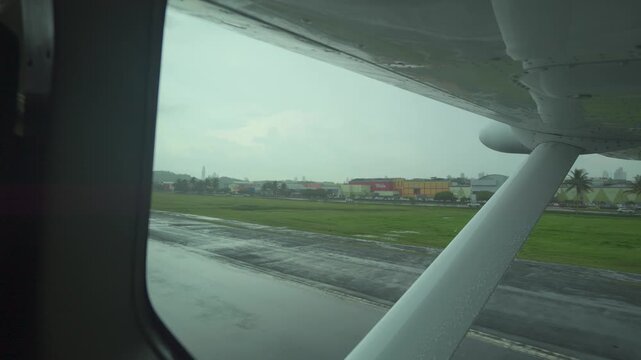 Raindrops streak the window pane.\nWet tarmac reflects distant structures.\nAirport surroundings blur in the downpour.\nClose-up shows raindrops on the window.\nIdeal for travel or weather-related con