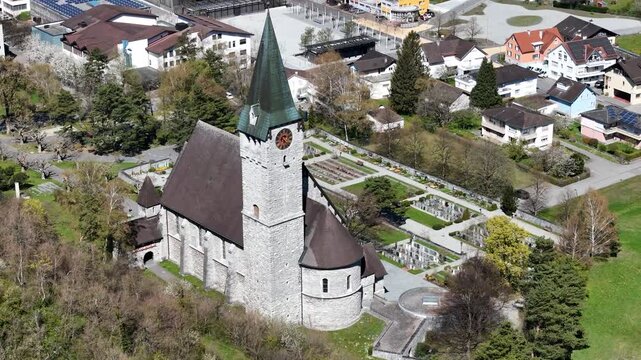 cemetery graveyard headstones rows garden pathways adjacent parish church clock face dark spire residential houses solar panels balzers liechtenstein drone footage close orbit heritage site