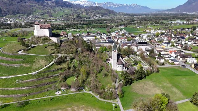 terraced vineyard slopes hillside castle round tower parish church spire snowcapped alpine peaks backdrop rhine valley farmland balzers town aerial panorama liechtenstein drone footage heritage site