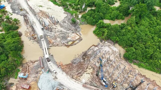 Aerial drone footage showcasing ongoing bridge foundation piling work, heavy machinery operation, and civil engineering progress at a remote river crossing construction site.