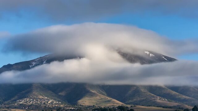 Cumulus clouds drift over a sunlit mountain peak during a clear day creating atmospheric perspective and natural beauty
