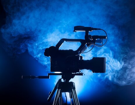 professional broadcast camera silhouetted against dramatic blue studio lights and atmospheric smoke ready for action