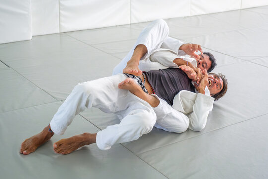 Two Brazilian jiu jitsu practitioners demonstrate rear choke defense from back control, with the front partner using both hands against the neck grip