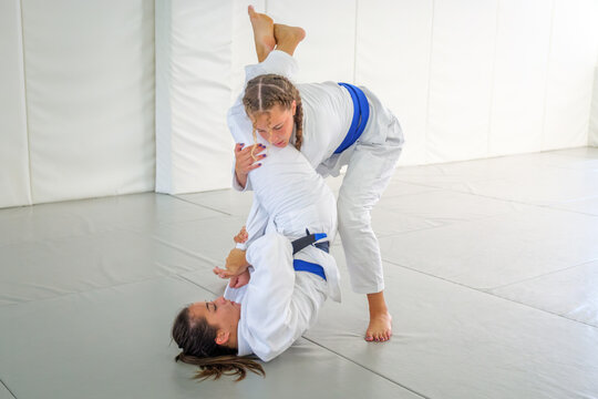 Two female athletes in white gis and blue belts practice a stack guard pass during Brazilian jiu jitsu training on an indoor martial arts mat