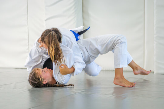Two female athletes practice brazilian jiu jitsu ground fighting on the tatami in a dojo. A student in a white gi and blue belt secures a pin on her partner
