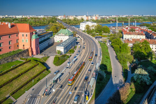 Elevated view of the W-Z Route below Warsaw Old Town with tram lines, road traffic, castle walls, city greenery, and historic buildings