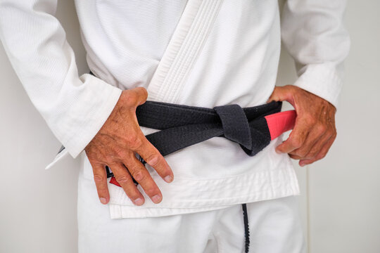 Close up of a tied Brazilian jiu jitsu black belt with a worn red rank bar over a white gi and the practitioner's hands near the knot