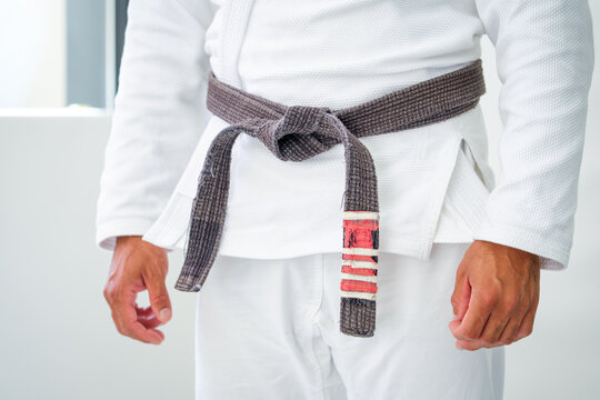 Side detail of a brown brazilian jiu jitsu belt knotted over a heavy white gi, the frayed rank bar hanging against the trousers