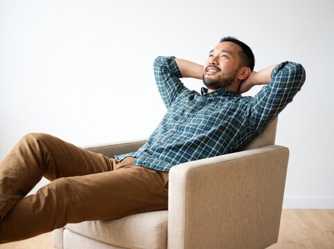 Happy Asian man relaxing in a comfortable armchair with hands behind his head, smiling and looking up in a bright minimalist room, enjoying a peaceful moment of rest, leisure, and deep satisfaction.
