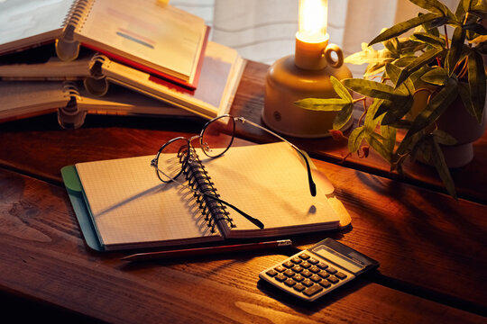 Notebooks, glasses, and a calculator on a wooden table with a warm light from a lamp in a cozy indoor setting