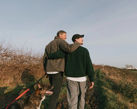 Two men walking a dog on a muddy field path, sharing a supportive embrace in the cold air for mens health awareness and mental wellness
